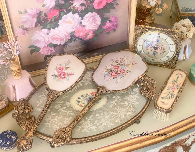 May include: Vintage vanity set featuring two hand mirrors and a brush with floral embroidery on a pale pink background. The set has ornate gold-colored metal handles and frames. A framed floral painting and a decorative clock are also visible.