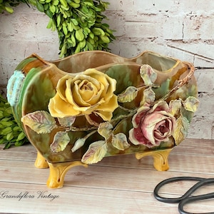 May include: A vintage ceramic planter with a floral design. The planter is rectangular with a green and brown glaze, featuring yellow and pink roses with detailed leaves. It has four yellow feet and the text "Grandiflora Vintage".
