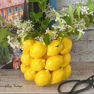 May include: A ceramic vase shaped like a cluster of bright yellow lemons, filled with green leafy branches and small white flowers. The vase has a glossy finish and a textured surface created by the individual lemon shapes. Black metal shears lie nearby.