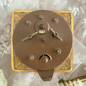 May include: Close-up of the back of a vintage clock. The clock's mechanism is visible, with a circular bronze-colored metal back and two silver-colored winding keys. The clock is set in a gold-colored, ornate rectangular frame. The text "MADE IN GT. BRITAIN" is visible.