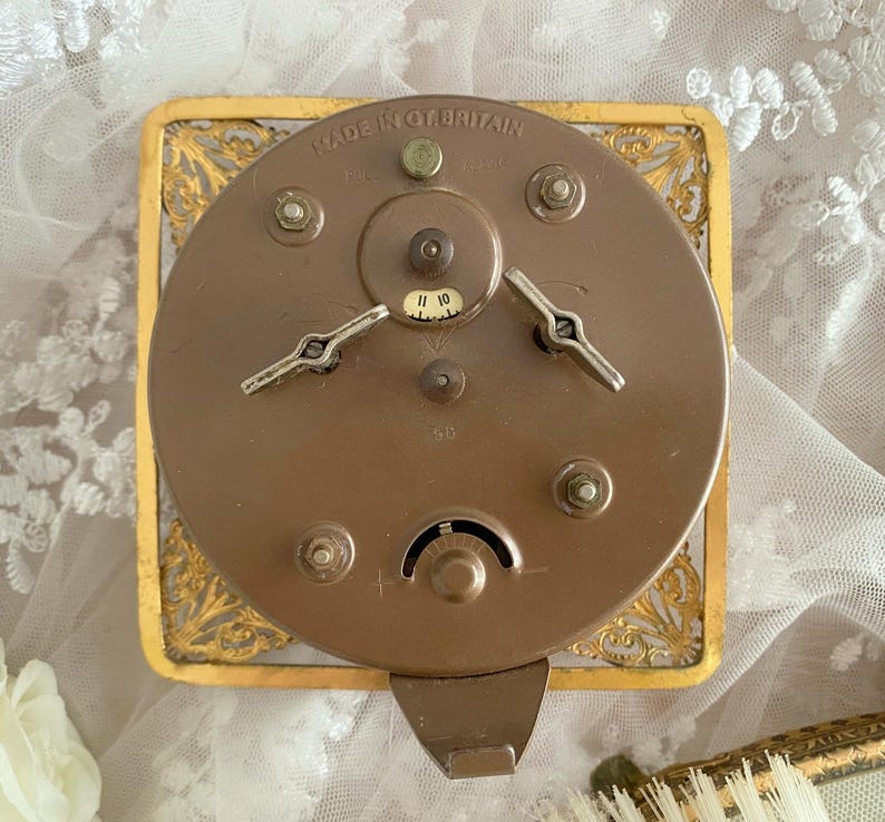 May include: Close-up of the back of a vintage clock. The clock's mechanism is visible, with a circular bronze-colored metal back and two silver-colored winding keys. The clock is set in a gold-colored, ornate rectangular frame. The text "MADE IN GT. BRITAIN" is visible.