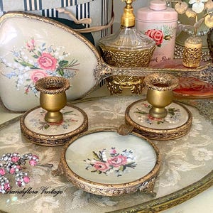 May include: An antique vanity set featuring a tray, two candle holders, and an ashtray. The set has a gold trim and floral embroidery in pink, blue, and white. The items are arranged on a decorative tray.