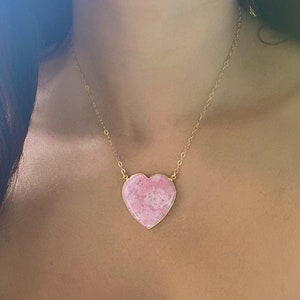 May include: A gold chain necklace with a large pink heart-shaped pendant. The pendant has a marbled pattern.