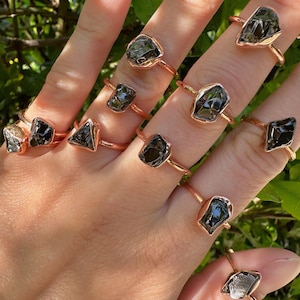 May include: A set of nine rings featuring raw, black gemstones set in rose gold. The rings are stacked on a hand, showcasing the unique shapes and sizes of the stones.