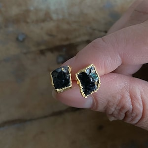 May include: A pair of black gemstone stud earrings with gold-colored trim. The earrings have a rough, natural appearance, with irregular edges. The gold setting frames the dark, textured stones, creating a contrast in color and texture. The earrings are small and delicate.