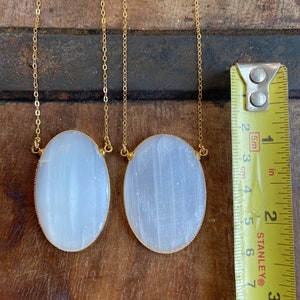 May include: Two oval-shaped selenite pendants on gold chains. The pendants are approximately 2 inches long and 1 inch wide. The selenite is a milky white color and has a smooth, polished finish.