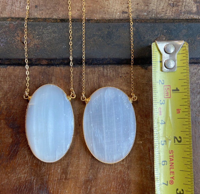 May include: Two oval-shaped selenite pendants on gold chains. The pendants are approximately 2 inches long and 1 inch wide. The selenite is a milky white color and has a smooth, polished finish.