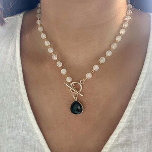 May include: A gold-toned necklace featuring a toggle clasp and a teardrop-shaped, dark green pendant. The necklace has a strand of small, round, white beads. The necklace is worn around the neck.