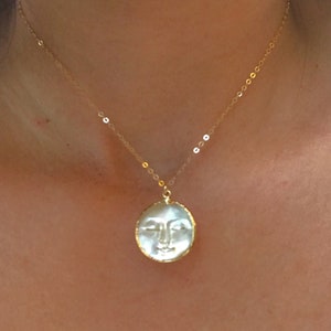 May include: A gold-colored necklace featuring a pendant with a carved face design. The pendant is round, with a white face and a gold-colored border. The necklace chain is delicate and gold-colored.