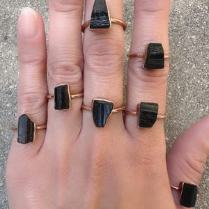 May include: A set of seven black tourmaline rings with rose gold bands. The rings feature raw, uncut black tourmaline stones of varying sizes and shapes.