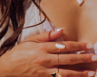 May include: A close-up of a hand wearing several gold rings with white and black gemstones. The rings are on the middle and ring fingers. The hand is resting on a white shirt.