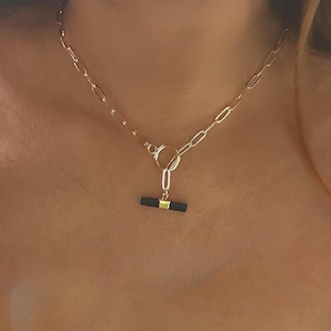 May include: Gold-toned chain necklace with a black and gold bar pendant. The necklace features a toggle clasp and a delicate chain design. The pendant is a dark black color with a gold accent.
