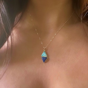 Pode incluir: Um colar de corrente de ouro com um pingente com duas pedras preciosas triangulares. A pedra superior é de cor azul claro e a pedra inferior é de cor azul escuro.