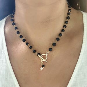 Puede incluir: Un delicado collar con cuentas negras ensartadas en una cadena dorada. El collar tiene un cierre de palanca y un pequeño dije con una cuenta negra y otra blanca. El collar se lleva alrededor del cuello.