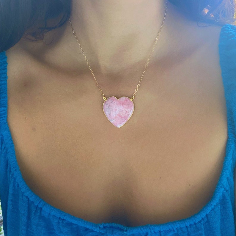 May include: A gold chain necklace with a large pink heart-shaped pendant. The pendant has a marbled pattern.
