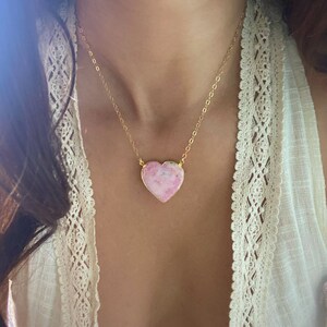 May include: A gold chain necklace with a pink heart-shaped pendant. The pendant has a marbled pattern and is made of natural stone.