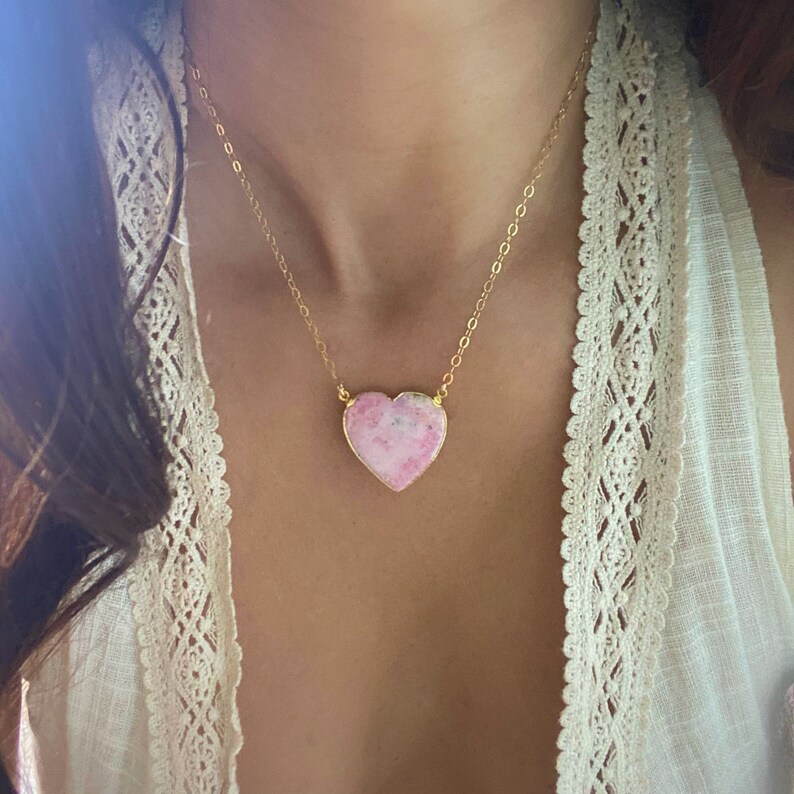 May include: A gold chain necklace with a pink heart-shaped pendant. The pendant has a marbled pattern and is made of natural stone.