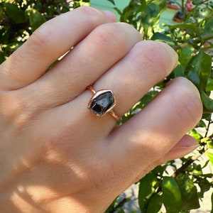 May include: A rose gold ring with a black, rough-cut gemstone. The ring is on a person's finger.