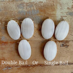 May include: Six oval-shaped selenite stones with gold-toned bails. Some stones have a single bail, while others have a double bail. The stones are arranged on a wooden surface. "Double Bail Or Single Bail" is written below the stones.