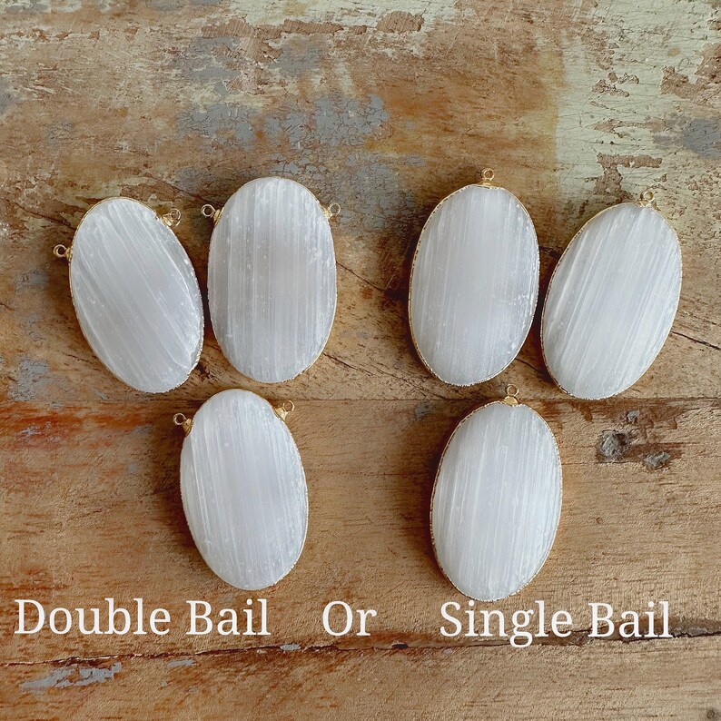 May include: Six oval-shaped selenite stones with gold-toned bails. Some stones have a single bail, while others have a double bail. The stones are arranged on a wooden surface. "Double Bail Or Single Bail" is written below the stones.