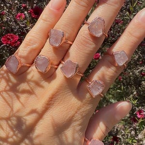 Op de afbeelding: Een set van zeven rozenkwarts ringen met ruwe kristallen die zijn gezet in roségouden banden. De ringen worden gedragen op een hand met een lichte huidskleur.