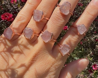 Puede incluir: Un conjunto de siete anillos de cuarzo rosa con piedras de cristal en bruto engastadas en bandas de oro rosa. Los anillos se llevan en una mano con un tono de piel claro.