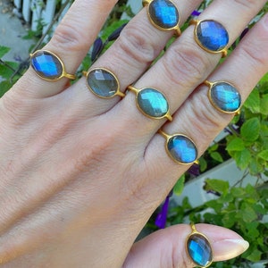 May include: A hand displays several gold-toned rings, each featuring an oval-shaped labradorite gemstone. The gemstones exhibit a play of color, with flashes of blue and green. The rings are set in a simple, elegant design.