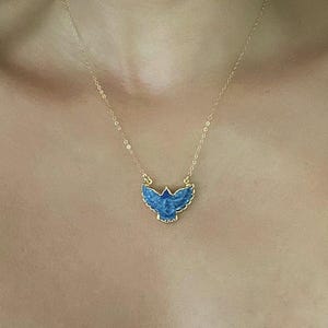 以下が含まれることがあります： 翼を広げた鳥の形をしたペンダントが付いたゴールドチェーンのネックレス。ペンダントは質感のある深い青色の素材で作られており、鳥の形を縁取るゴールドトーンの金属製の枠がチェーンにつながっています。