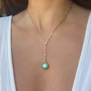 May include: Gold-toned Y-shaped necklace with a teardrop-shaped pendant. The pendant features a green and blue iridescent gemstone. The necklace is made of linked chain and is worn around the neck.