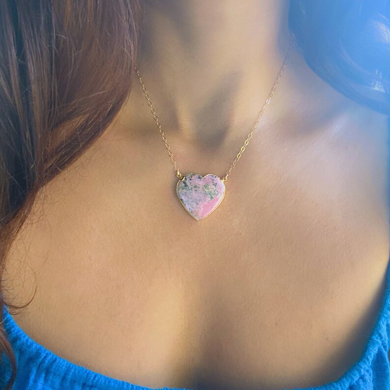 May include: A gold chain necklace with a large pink heart-shaped pendant. The pendant is made of a pink and green stone with a marbled pattern.