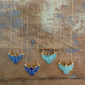 May include: Four gold necklaces with bird-shaped pendants. The pendants have gold edging and are in two colours: deep blue and light teal. The necklaces hang against a wooden background. The chains are of a fine design.