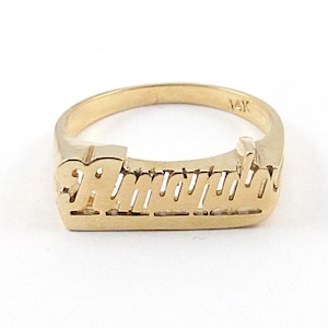 May include: A gold ring featuring a nameplate design with the script text "Amara" centered on the front. The band is smooth and polished, and the interior of the band is clearly stamped with the hallmark "14K" indicating the gold purity.