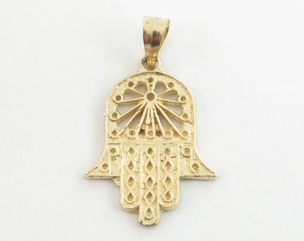 May include: A gold-colored pendant in the shape of a hamsa hand, a symbol of protection and good luck. The pendant is intricately detailed with a circular design in the center and a pattern of small circles around the edges.
