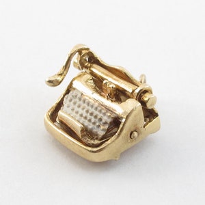 May include: A gold-coloured charm in the shape of a vintage typewriter. The charm is detailed with a small carriage return lever and a visible typewriter key.