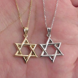 Puede incluir: Dos collares con colgantes de estrella de David. Uno es dorado, el otro plateado. Ambos colgantes cuelgan de delicadas cadenas. Los collares se muestran sobre un fondo color piel.