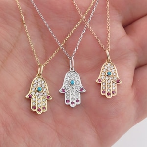 May include: Three gold necklaces with hamsa hand pendants. Each pendant is adorned with small white stones and a turquoise blue stone in the centre. The hamsa hands are outlined with small red stones.