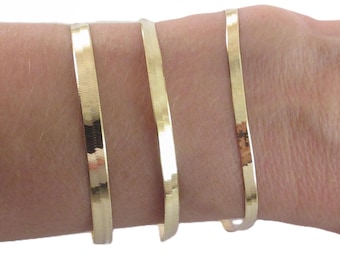 May include: Three gold-colored, thin, flat bracelets are worn on a wrist. The bracelets are made of a shiny, metallic material. The bracelets are simple and elegant, suitable for everyday wear.