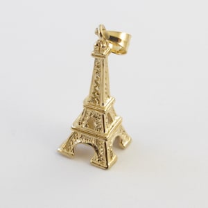 K&ouml;nnte beinhalten: Ein goldfarbener Eiffelturm-Anh&auml;nger. Der Anh&auml;nger ist eine Miniatur-Nachbildung des ber&uuml;hmten Pariser Wahrzeichens mit aufwendigen architektonischen Details. Der Anh&auml;nger hat oben eine Schlaufe zum Befestigen an einer Kette. Der Hintergrund ist wei&szlig;.