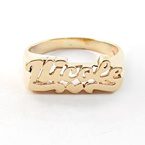 May include: A gold-toned metal ring featuring the name Nicole in a cursive script font. The design includes a small heart shape integrated into the bottom center of the lettering, with a smooth, polished finish across the entire surface of the band.