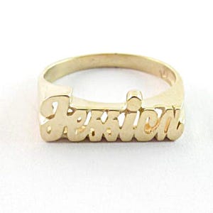 May include: A gold-toned metal ring featuring the name Jessica in a cursive script font. The letters are integrated into the band, creating a solid, raised nameplate design that spans the front of the ring with a polished, reflective surface finish.