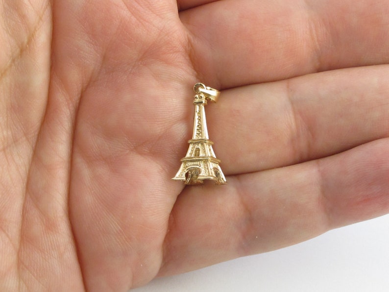 K&ouml;nnte beinhalten: Ein goldfarbener Eiffelturm-Anh&auml;nger. Der Anh&auml;nger ist eine detaillierte Miniaturreplik des ber&uuml;hmten Pariser Wahrzeichens. Der Anh&auml;nger ist an einer kleinen Schlaufe oben befestigt, bereit, an einer Halskette oder einem Armband befestigt zu werden. Der Anh&auml;nger wird in einer Hand gehalten.