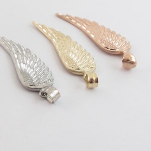Op de afbeelding: Drie gouden engelenvleugelcharms, een in zilver, een in goud en een in ros&eacute; goud. Elke charm heeft een gedetailleerd vleugelontwerp en een lus om aan een ketting of armband te bevestigen.