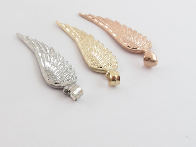 Op de afbeelding: Drie gouden engelenvleugelcharms, een in zilver, een in goud en een in ros&eacute; goud. Elke charm heeft een gedetailleerd vleugelontwerp en een lus om aan een ketting of armband te bevestigen.