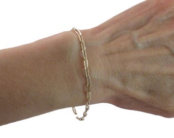 May include: A gold-colored paperclip chain bracelet. The bracelet is made of small, rectangular links and is fastened with a lobster clasp. The bracelet is shown on a person's wrist.