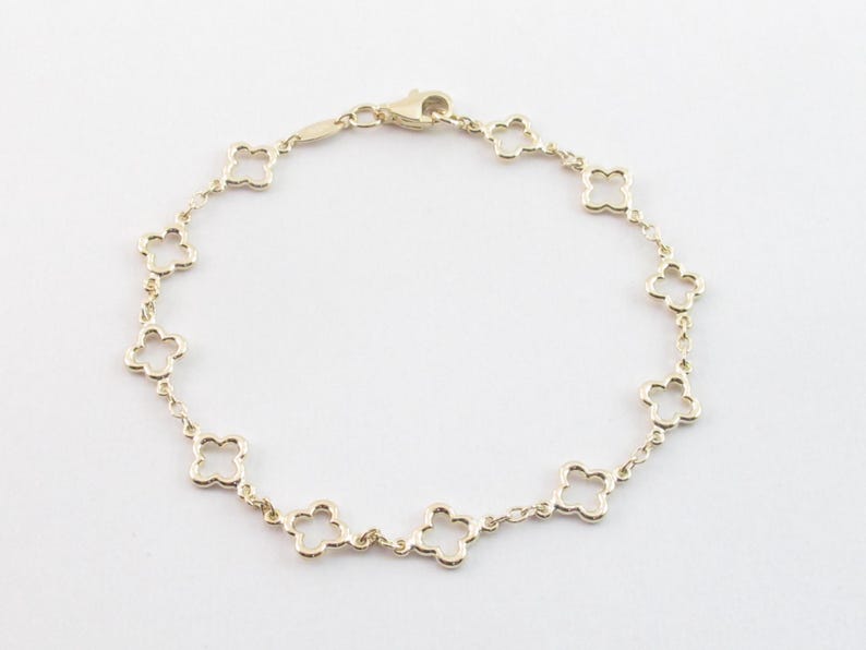 May include: A gold-colored bracelet featuring a delicate clover design. The bracelet is composed of multiple clover-shaped links connected by small chain segments, with a clasp closure. The jewelry is set against a white background.