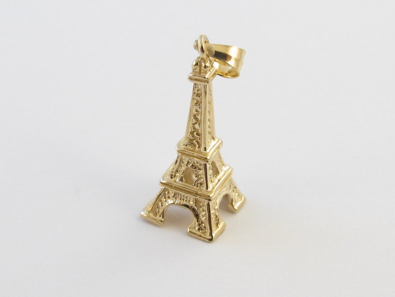 K&ouml;nnte beinhalten: Ein goldfarbener Eiffelturm-Anh&auml;nger. Der Anh&auml;nger ist eine Miniatur-Nachbildung des ber&uuml;hmten Pariser Wahrzeichens mit aufwendigen architektonischen Details. Der Anh&auml;nger hat oben eine Schlaufe zum Befestigen an einer Kette. Der Hintergrund ist wei&szlig;.