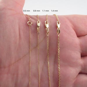 May include: Four gold-colored chain necklaces of varying thicknesses (0.6 mm, 0.8 mm, 1.1 mm, and 1.4 mm) are displayed. Each chain has a clasp. The chains are held in a person's open hand, showcasing the different chain styles.