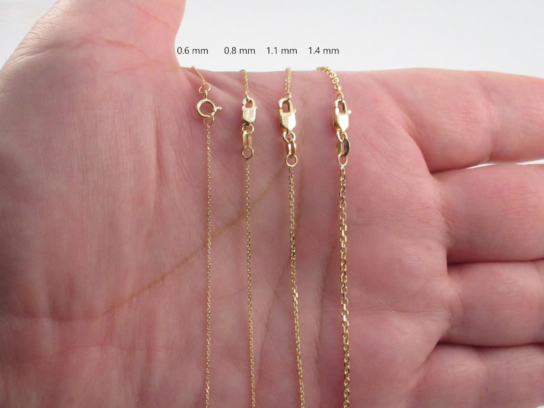 May include: Four gold-colored chain necklaces of varying thicknesses (0.6 mm, 0.8 mm, 1.1 mm, and 1.4 mm) are displayed. Each chain has a clasp. The chains are held in a person's open hand, showcasing the different chain styles.