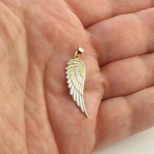 Op de afbeelding: Een gouden en zilveren engelenvleugel hanger. De vleugel is gedetailleerd met getextureerde lijnen en heeft een lus aan de bovenkant om aan een ketting te bevestigen.