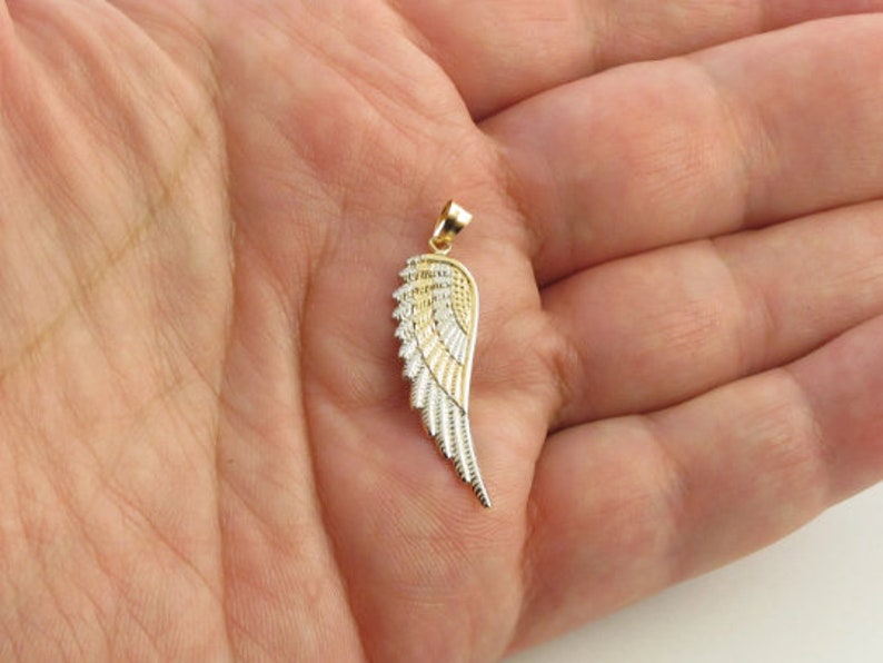 Op de afbeelding: Een gouden en zilveren engelenvleugel hanger. De vleugel is gedetailleerd met getextureerde lijnen en heeft een lus aan de bovenkant om aan een ketting te bevestigen.